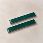 Two glossy dark green rectangular ceramic tiles with silver metal backings, displayed diagonally on a textured beige background.