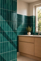 A modern kitchen corner featuring glossy emerald green vertical subway tiles on the wall, light wood cabinets, a white countertop, and a window with natural light.
