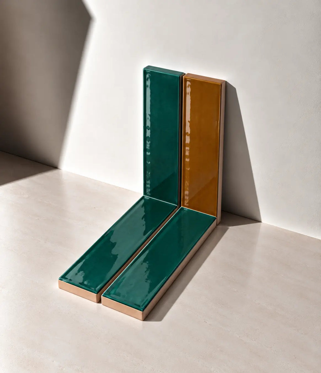 Glossy emerald green and cognac handmade ceramic tiles arranged against a white wall in natural sunlight.