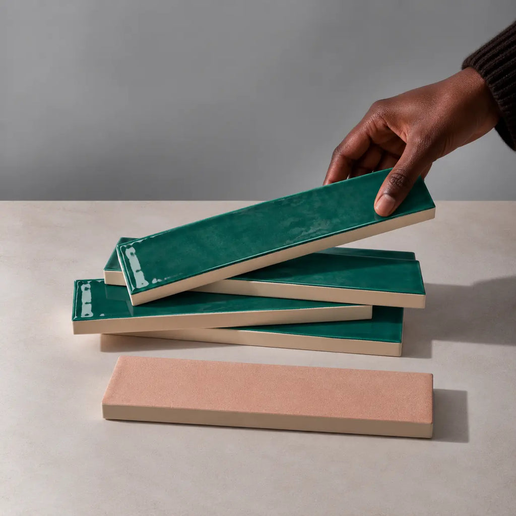 Hand holding a glossy emerald green rectangular ceramic tile above a stack of handmade tiles on a neutral stone surface.