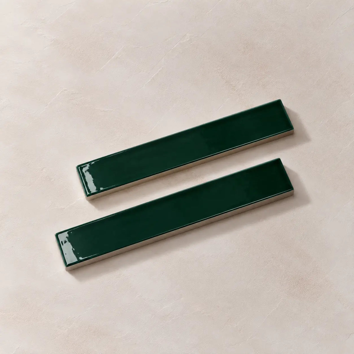 Two glossy dark green rectangular ceramic tiles with silver metallic edges, arranged diagonally on a textured beige background.
