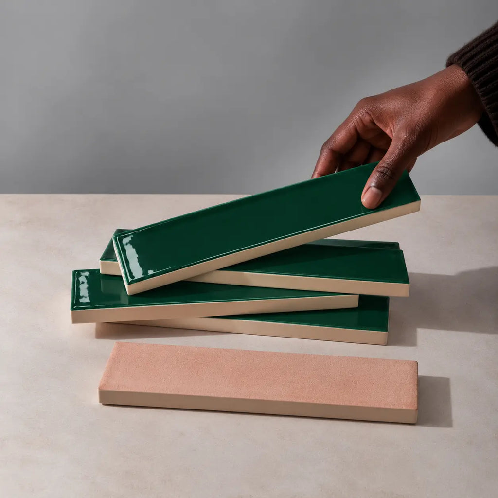 Hand holding a glossy forest green rectangular ceramic tile above a stack of handmade tiles on a neutral stone surface.