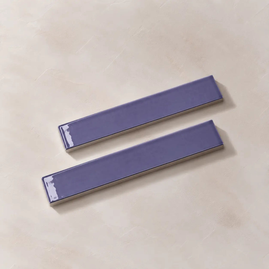 Two glossy lilac rectangular ceramic tiles with a textured beige base, arranged diagonally on a light beige textured surface.