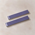Two glossy lilac rectangular ceramic tiles with a textured beige base, arranged diagonally on a light beige textured surface.