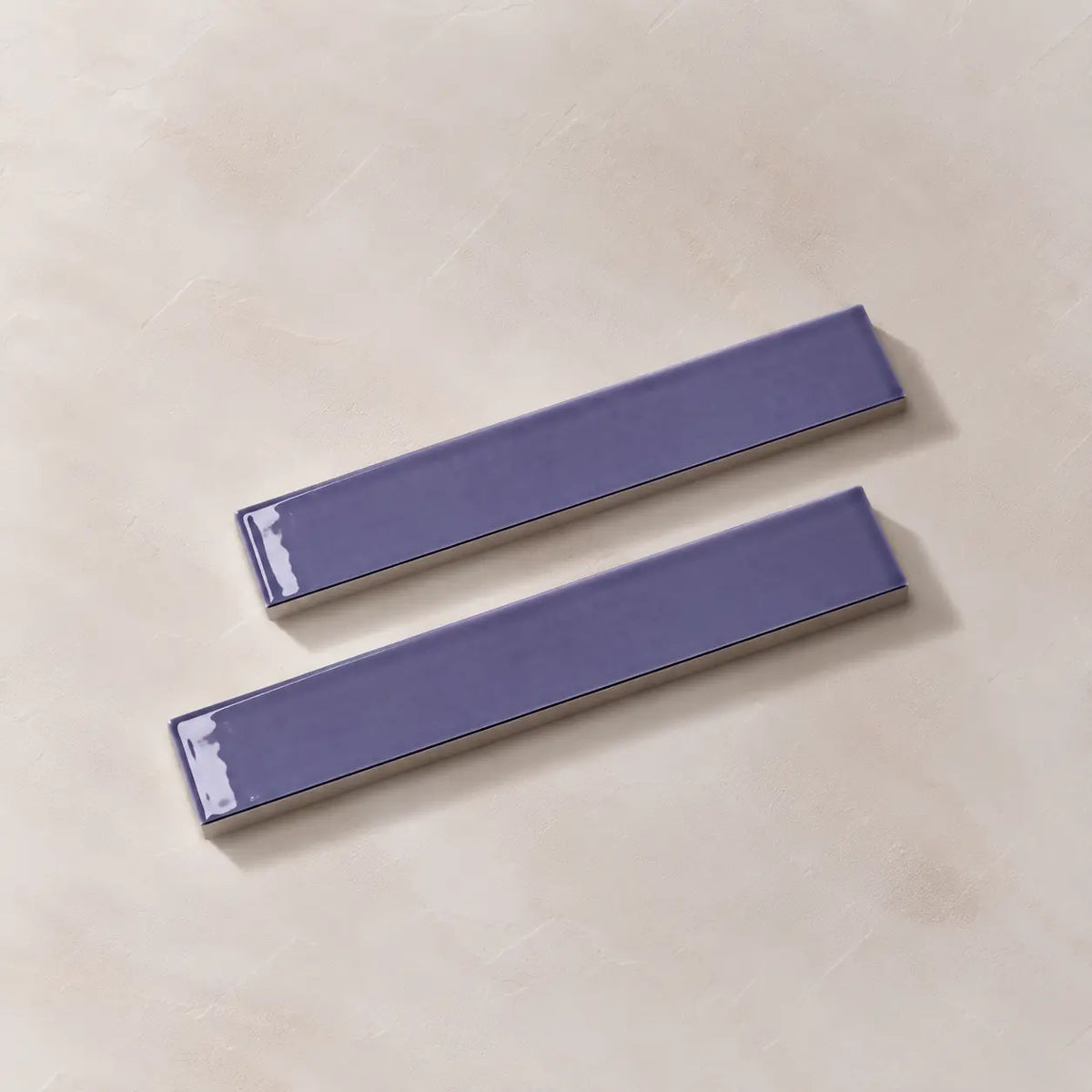 Two glossy lilac rectangular ceramic tiles with a textured beige base, arranged diagonally on a light beige textured surface.