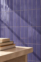 Purple glossy rectangular wall tiles in lilac colour, arranged in a grid pattern, with a beige stone ledge in the foreground holding a stack of books.