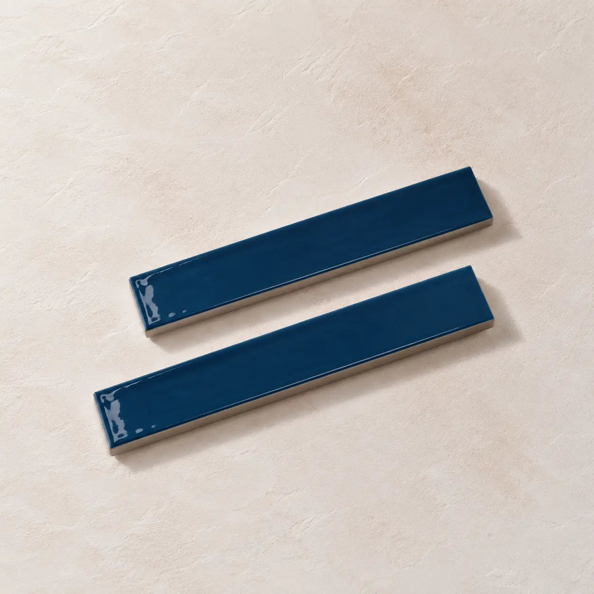 Two glossy dark blue rectangular ceramic tiles with a beveled edge, displayed diagonally on a textured beige background.