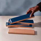 Hand holding a glossy mediterranean blue rectangular ceramic tile above a stack of handmade tiles on a neutral stone surface.