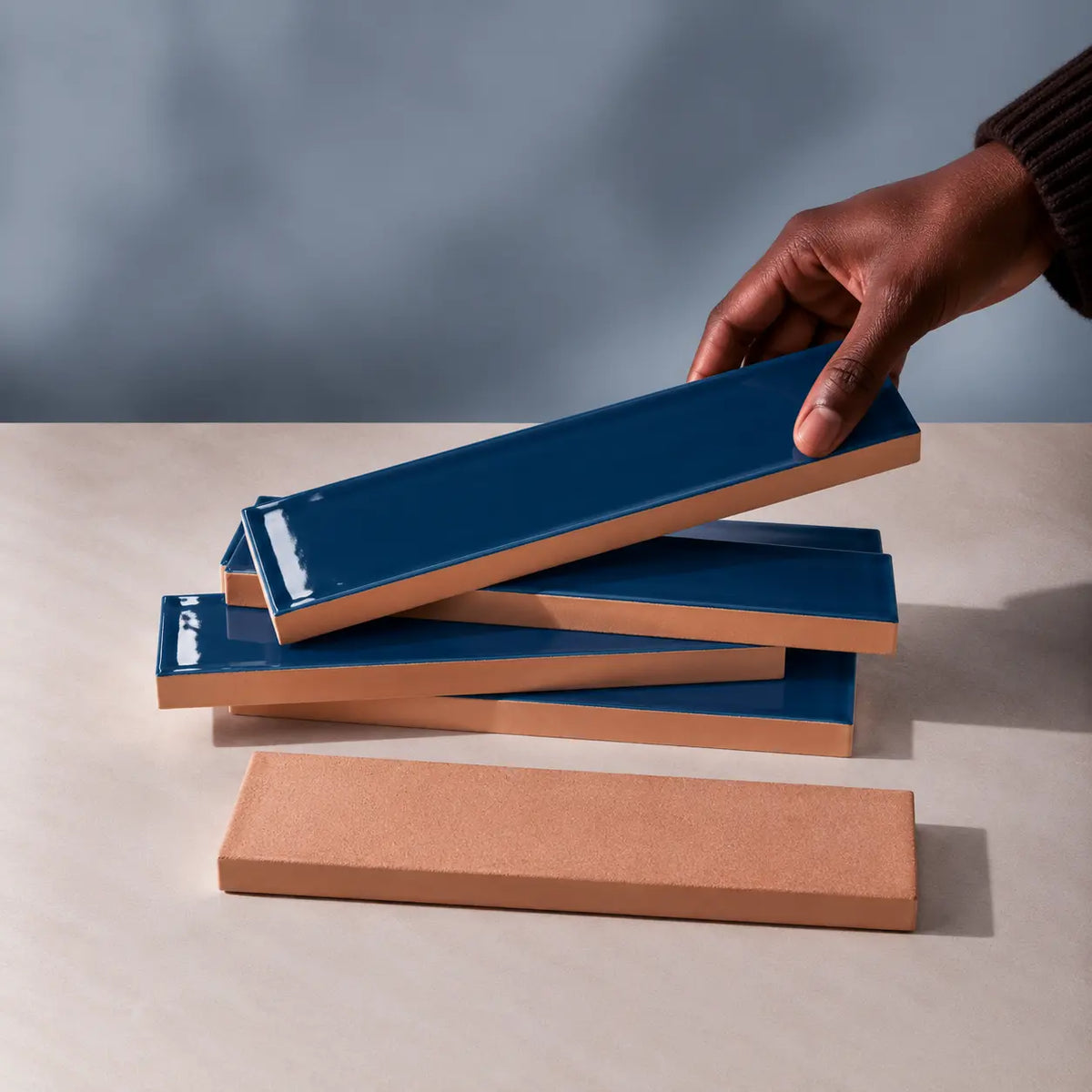 Hand holding a glossy mediterranean blue rectangular ceramic tile above a stack of handmade tiles on a neutral stone surface.