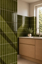 Green glossy rectangular wall tiles in moss green installed in a kitchen setting, paired with light wood cabinets and a white countertop under a window.