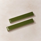 Two glossy olive green rectangular ceramic tiles with a slight bevelled edge, displayed diagonally on a textured beige background.