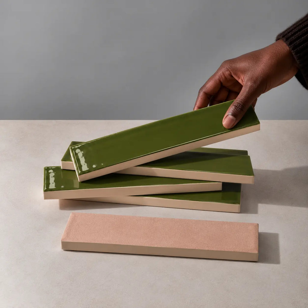 Hand holding a glossy olive green rectangular ceramic tile above a stack of handmade tiles on a neutral stone surface.