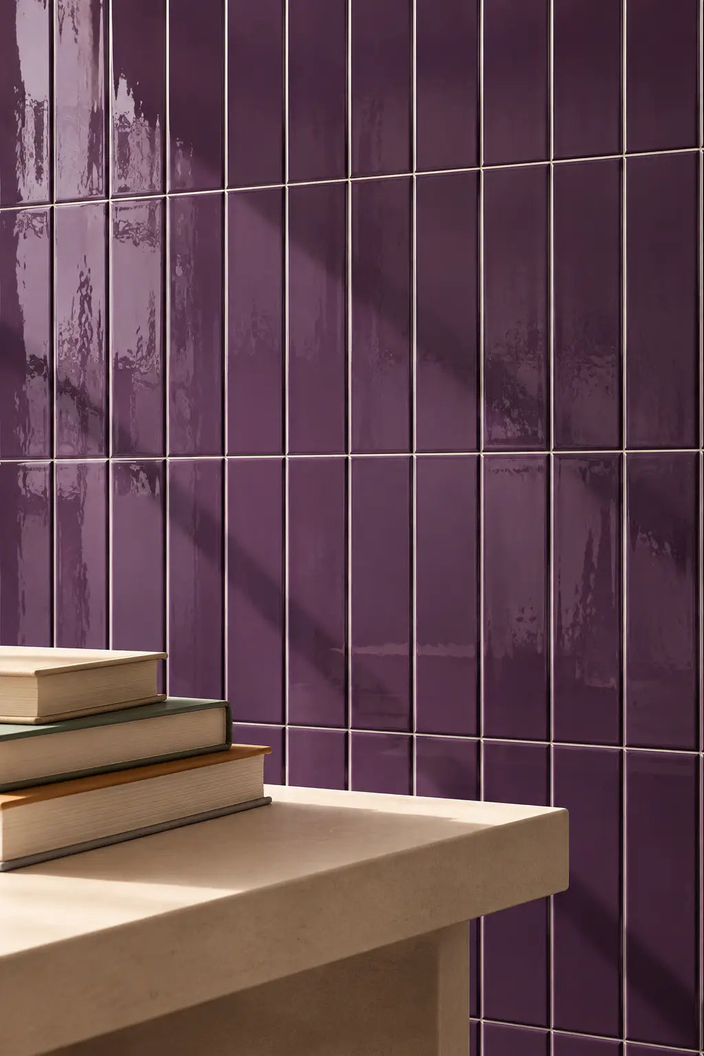 Purple glossy rectangular wall tiles in plum colour, arranged in a grid pattern, with a light beige stone ledge in the foreground holding a stack of books.
