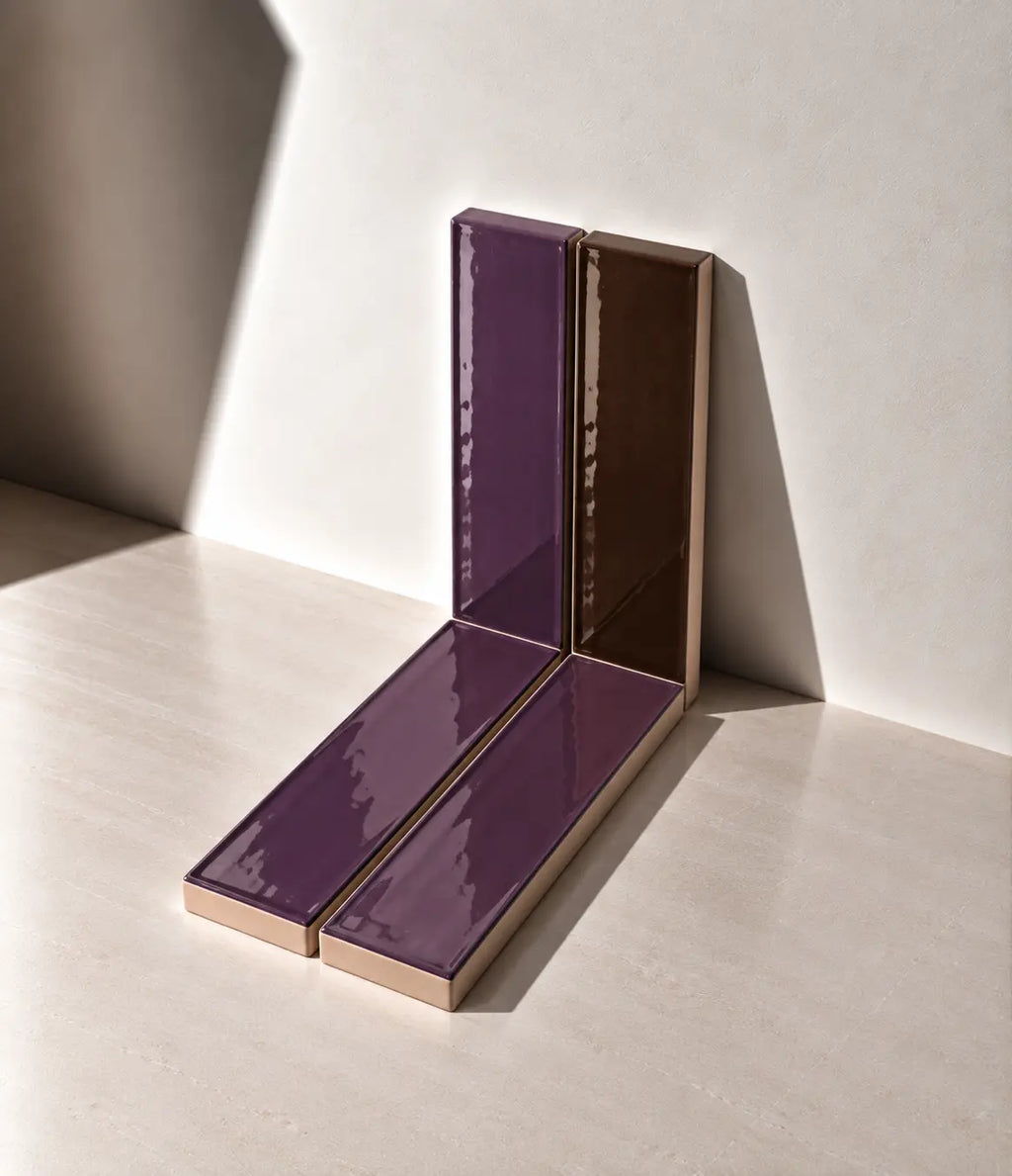 Glossy plum and chocolate brown handmade ceramic tiles arranged against a white wall in natural sunlight.