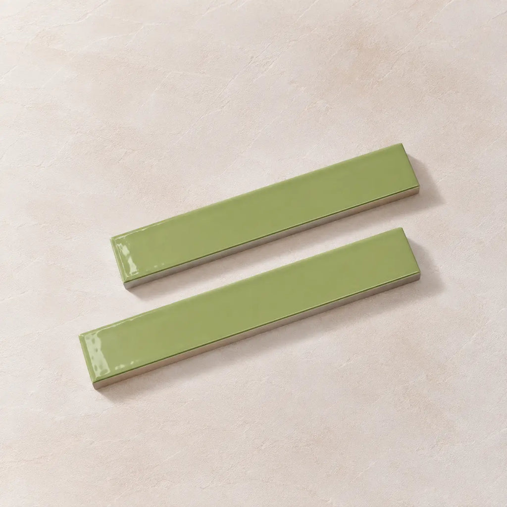 Two glossy rectangular ceramic tiles in a light sage green color, arranged diagonally on a textured beige background.