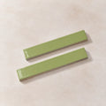Two glossy rectangular ceramic tiles in a light sage green color, arranged diagonally on a textured beige background.