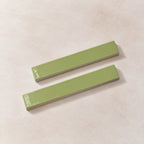 Two glossy rectangular ceramic tiles in a light sage green color, arranged diagonally on a textured beige background.