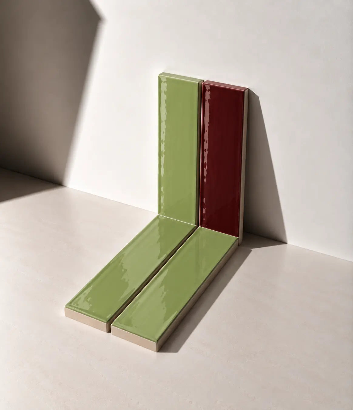 Glossy Sage Green and Burgundy Red handmade ceramic tiles arranged against a white wall in natural sunlight.
