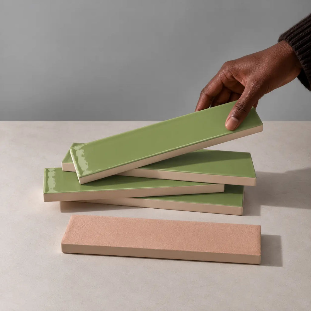 Hand holding a glossy sage green rectangular ceramic tile above a stack of handmade tiles on a neutral stone surface.