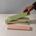 Hand holding a glossy sage green rectangular ceramic tile above a stack of handmade tiles on a neutral stone surface.