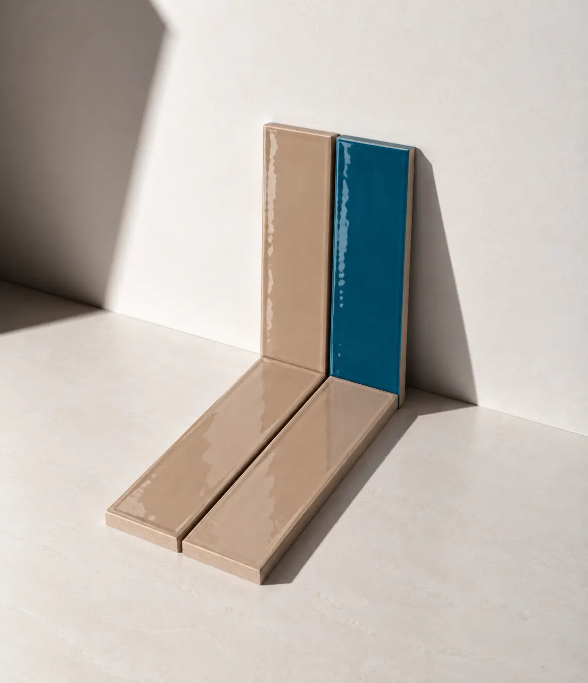 Two glossy ceramic tiles, one in sand beige and one in teal blue, arranged in a V-shape against a minimalist off-white wall and floor with dramatic shadows.