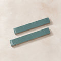Two rectangular glossy teal ceramic subway tiles on a textured beige background.