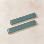 Two rectangular glossy teal ceramic subway tiles on a textured beige background.
