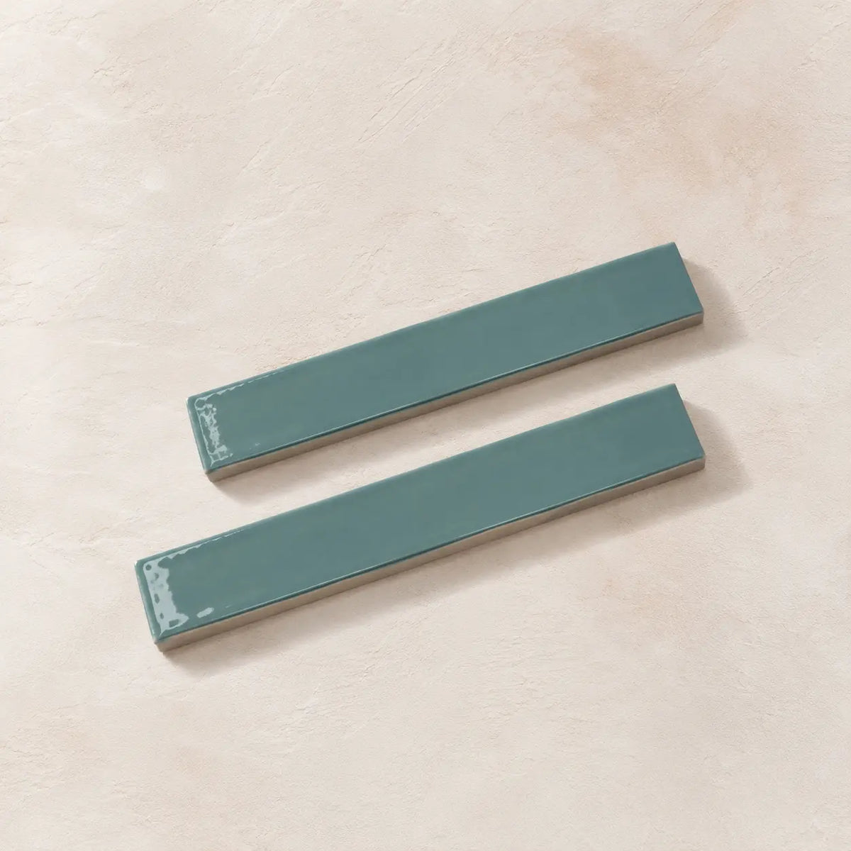 Two rectangular glossy teal ceramic subway tiles on a textured beige background.