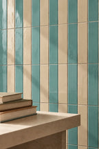 Vertical striped ceramic wall tiles in teal and cream, shown behind a light stone ledge with stacked books in the foreground.