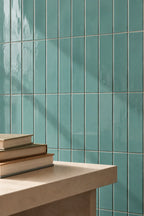 Glossy vertical teal ceramic tiles on a wall behind a light beige stone counter with stacked books in the foreground.