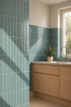 A modern kitchen corner with glossy vertical teal-blue tiles, a light wood cabinet, a white countertop, and a small potted plant near a window.