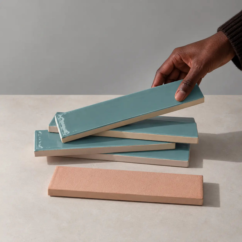 Hand holding a glossy blue sky rectangular ceramic tile above a stack of handmade tiles on a neutral stone surface.