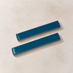 Two glossy teal rectangular ceramic tiles with rounded corners, displayed diagonally on a textured beige background.