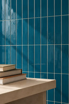 Glossy vertical teal-blue ceramic tiles with thin white grout lines, shown behind a light beige stone ledge with stacked books in the foreground.