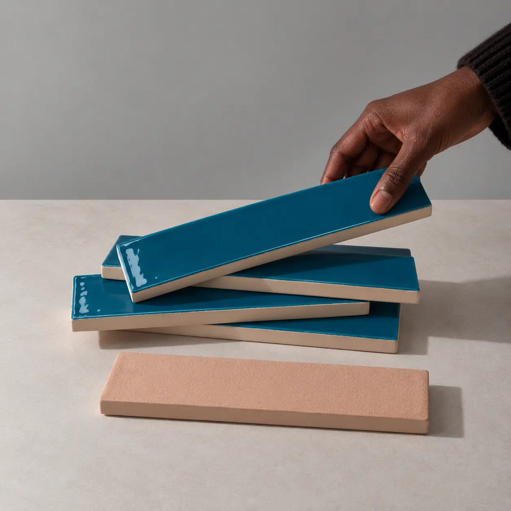 Hand holding a glossy teal blue rectangular ceramic tile above a stack of handmade tiles on a neutral stone surface.