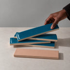 Hand holding a glossy teal blue rectangular ceramic tile above a stack of handmade tiles on a neutral stone surface.