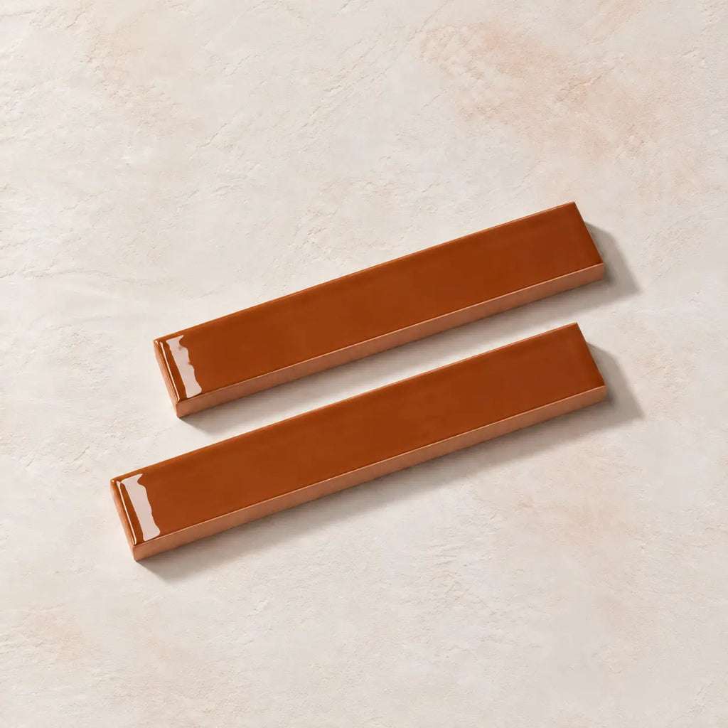 Two glossy terracotta orange rectangular ceramic tiles with a textured beige base, arranged diagonally on a light beige textured surface.