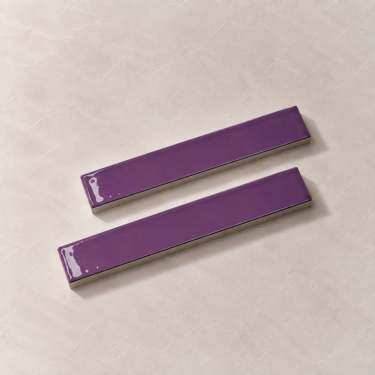 Two glossy purple rectangular ceramic tiles with silver metallic edges, displayed diagonally on a light beige textured background.