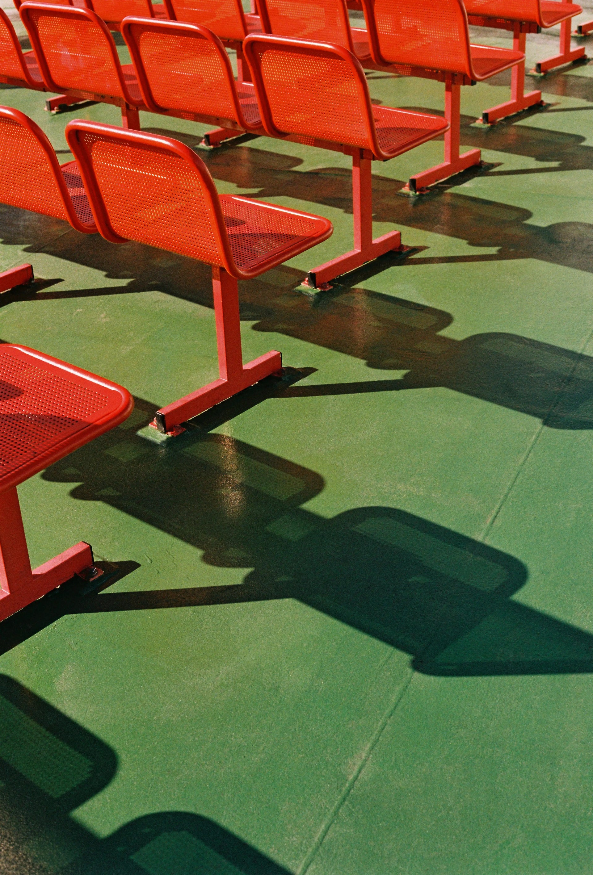 Red metal stadium seating rows on a green floor with strong shadows.