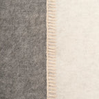 Close-up of a throw blanket with a vertical seam, showing a dark gray felt-like side and an off-white woven side with cream fringe.
