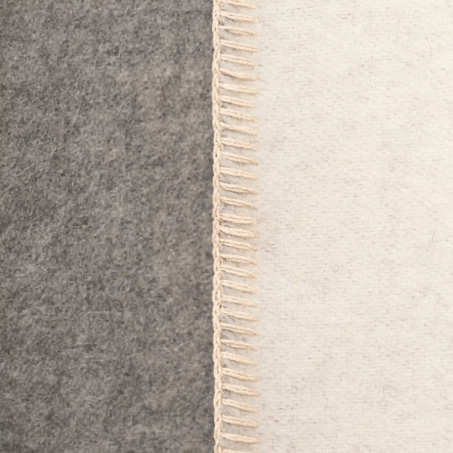Close-up of a throw blanket with a vertical seam, showing a dark gray felt-like side and an off-white woven side with cream fringe.