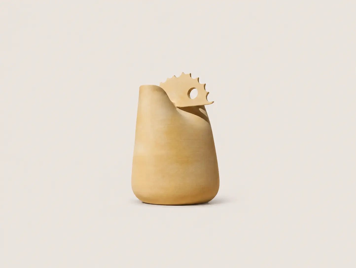 A sculptural ceramic pitcher or vase in a warm sandy beige color, featuring a wide flared rim and a decorative serrated tab on top with a circular cutout, set against a solid light beige background.