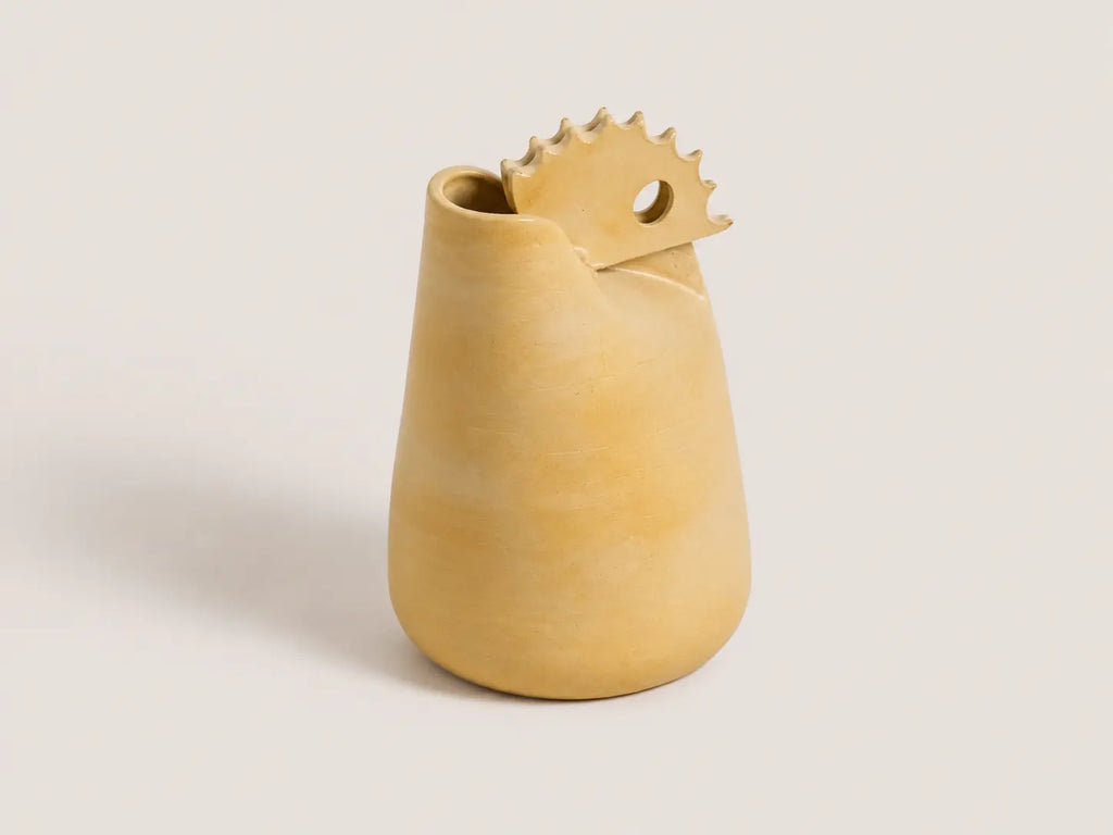 A small sculptural ceramic vase in a warm sandy beige color, featuring a tapered body and a spout-like top with a circular cutout, set against a solid light gray background.