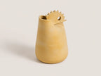 A small sculptural ceramic vase in a warm sandy beige color, featuring a tapered body and a spout-like top with a circular cutout, set against a solid light gray background.