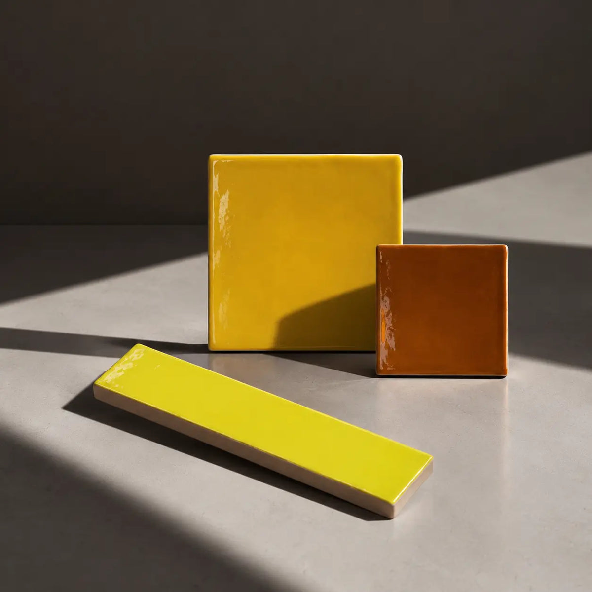 Three glossy ceramic tiles in yellow and orange-brown shades arranged on a gray surface with dramatic diagonal sunlight and shadows.