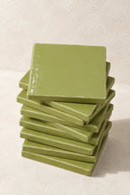 A stack of glossy square ceramic coasters in a muted sage green color, set against a textured off-white background.
