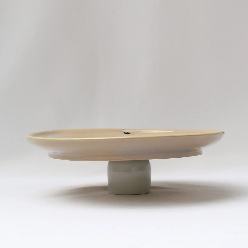 Minimal side view of a cream ceramic dinner plate with a raised pedestal-style base against a neutral background.