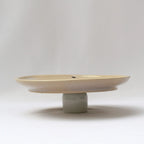 Minimal side view of a cream ceramic dinner plate with a raised pedestal-style base against a neutral background.