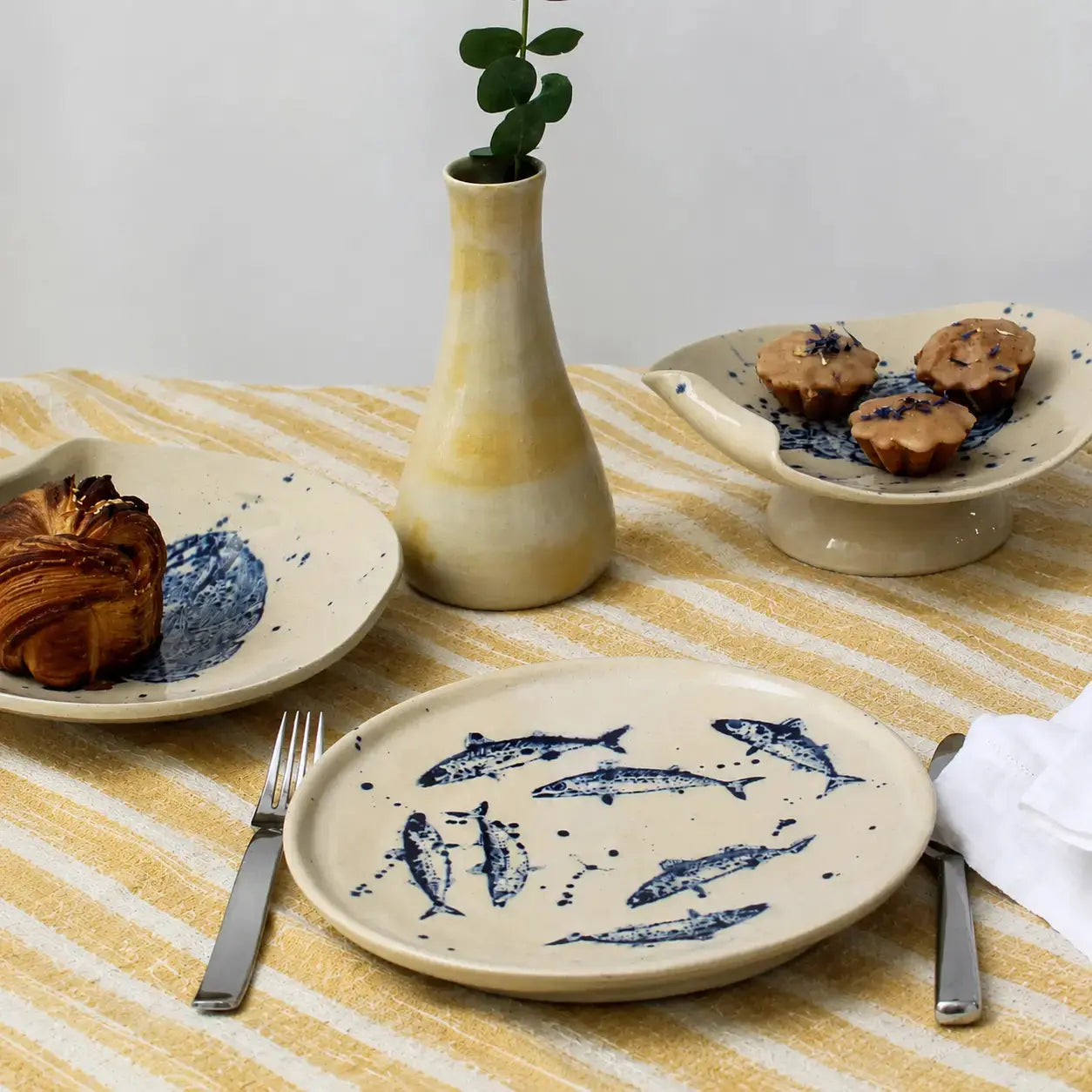 Ceramic plates with fish design on a striped tablecloth, accompanied by a vase and pastries.