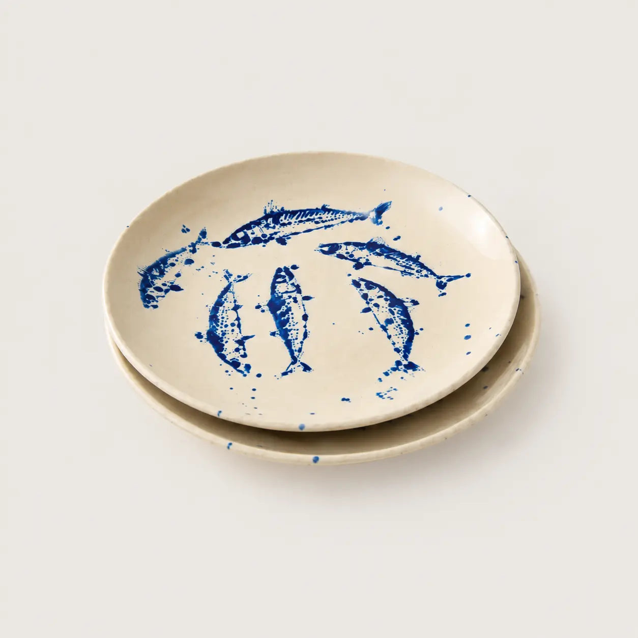 A set of two round ceramic plates in a warm cream color, decorated with blue ink-style illustrations of fish and blue paint splatters, set against a plain light gray background.